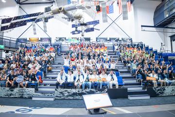 Teilnehmerinnen und Teilnehmer des internationalen CanSat-Finales jubeln auf der Tribüne im ESA-Forschungszentrum ESTEC unter Modellen von Satelliten und Raumstation